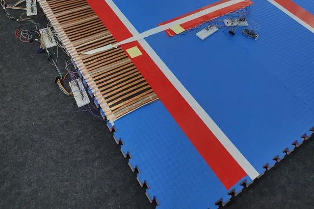 A competition mat lifted at one corner to reveal rows of copper-taped pressure-sensing strips wired into breadboards.